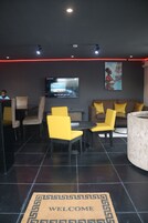 A modern lounge area with a 'Welcome' mat, seating arrangements, a flat-screen TV, and a decorative wall piece.