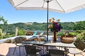 Un patio al aire libre con una mesa dispuesta para dos personas, rodeado de sillones de mimbre, un paraguas grande y un jarrón con flores.