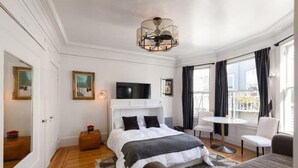 Quarto de hotel com cama, televisão, mesa pequena e cadeira. Tem uma janela com cortinas e vista para o exterior.