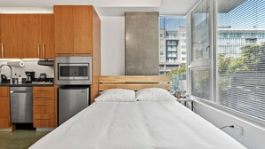 A modern hotel room with a wooden bed, a kitchenette with a microwave and refrigerator, and a large window with blinds. - Studio apt walkable to Oracle Park (San Francisco)