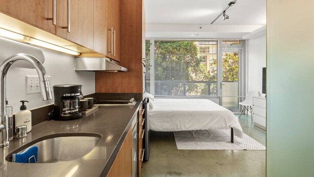 A modern kitchen with a coffee maker, a sink, and wooden cabinets. There is a bed with a white cover and a view of greenery through large windows. - Studio apt walkable to Oracle Park (San Francisco)