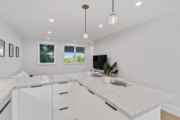 A modern kitchen with white cabinetry, a marble countertop, pendant lighting, and a large window.