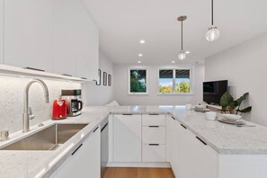 A modern kitchen with white cabinetry, a stainless steel sink, a red coffee maker, and a large window providing a view of greenery outside. - Modern 1BR Noe Valley Apt with laundry & backyard (San Francisco)