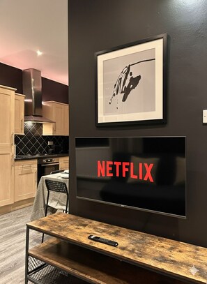 Eine moderne Küche mit Esstisch und Stühlen, einem an der Wand montierten Fernseher mit Netflix-Logo und einem gerahmten Kunstwerk darüber.