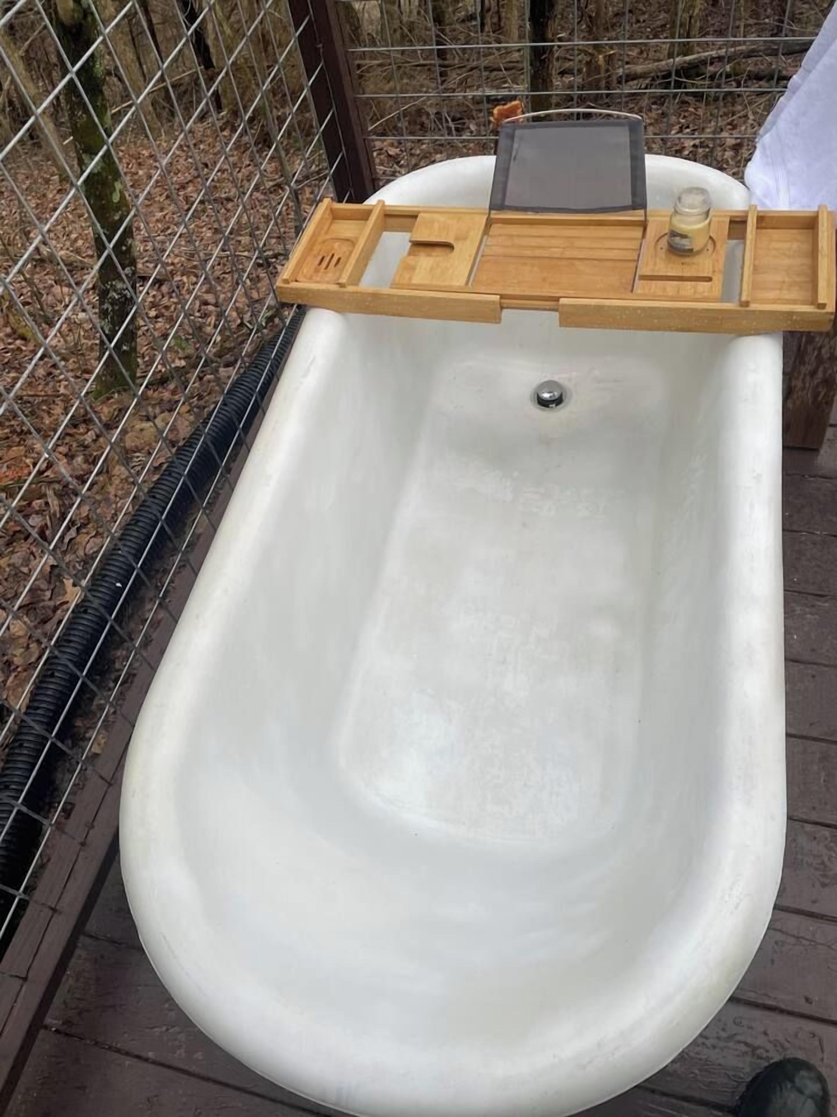 A white outdoor bathtub with a wooden tray on top, surrounded by a metal fence.