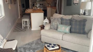 A compact living space with a kitchenette, a sofa with patterned cushions, a wicker coffee table, and a woven rug.