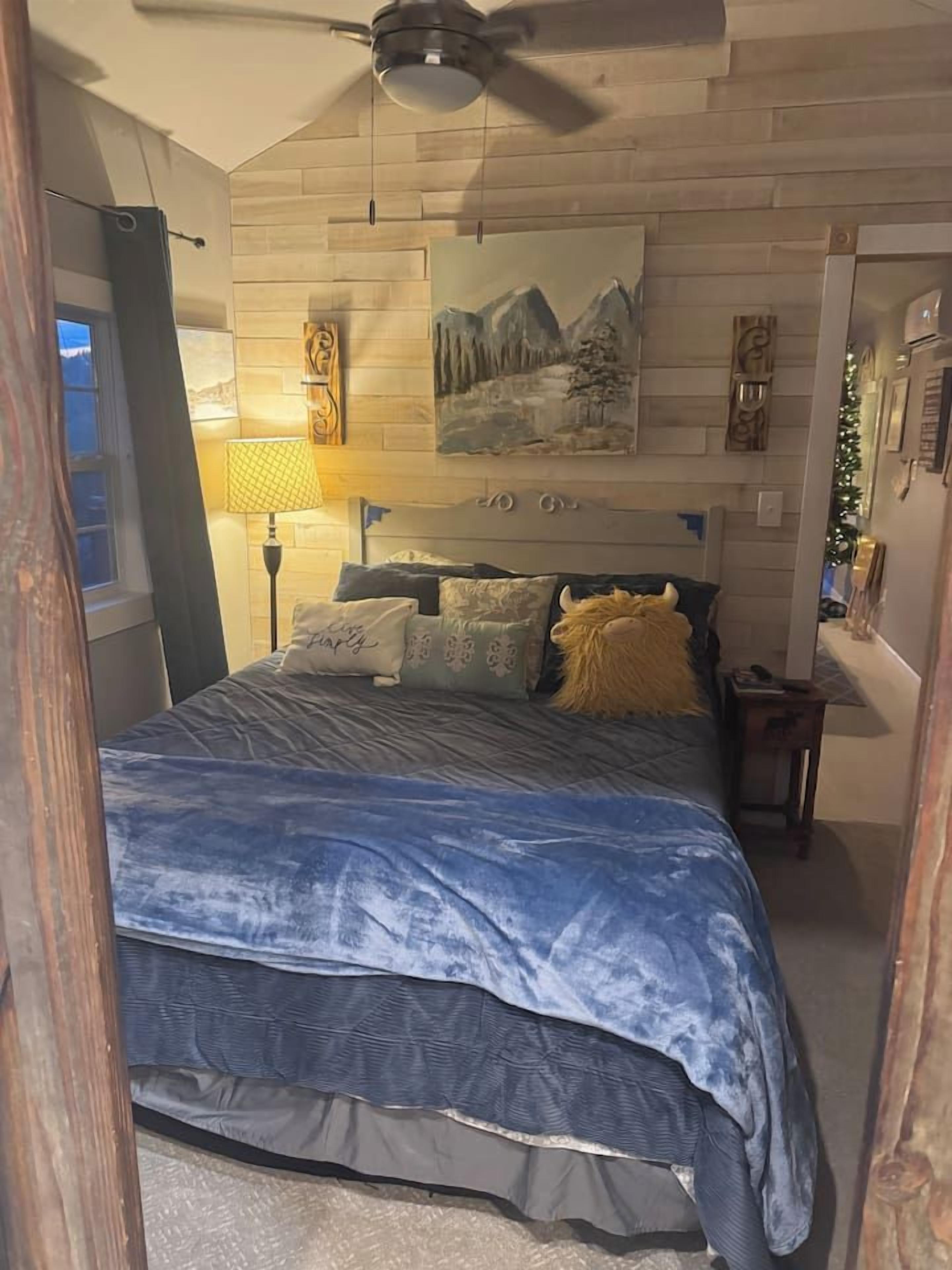 A bedroom with a bed, a ceiling fan, a painting, and a nightstand.