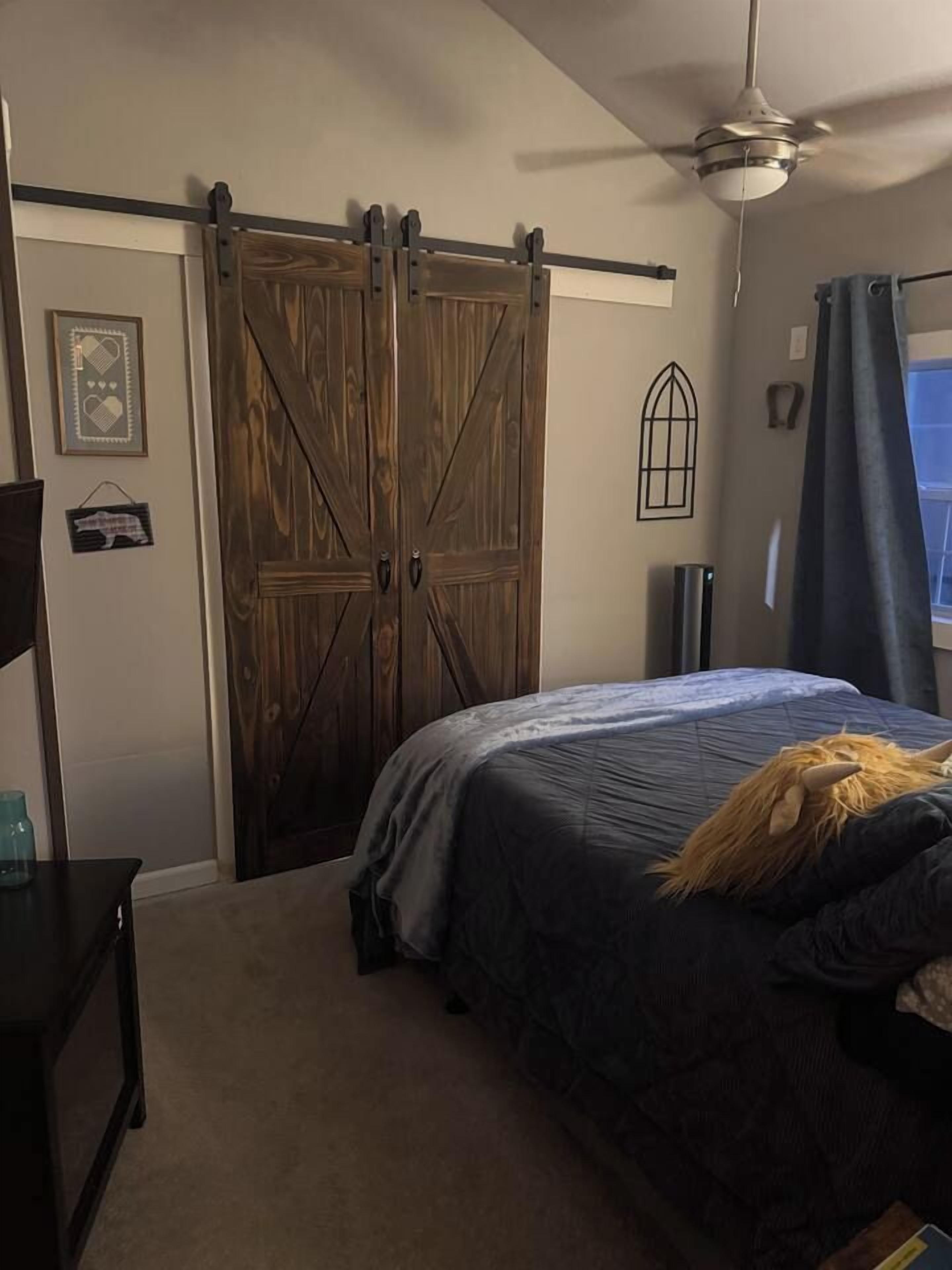 A bedroom with a bed, a ceiling fan, a sliding barn door, and a window with curtains.
