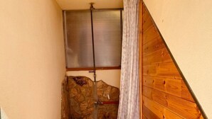 Ein schmaler, abgeschlossener Raum mit Steinwand, Holzvertäfelung, einer Metalttür und einem Fenster mit Rollläden.