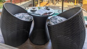 Ein Balkon mit Korbstühlen, einem kleinen Tisch mit Tasse und Untertasse sowie Blick auf einen Poolbereich mit Palmen und Gebäuden im Hintergrund.