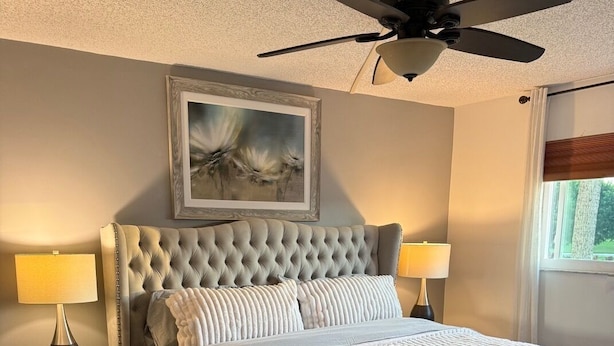 Een slaapkamer met een gestoffeerd hoofdeinde, een groot bed met een geborduurde deken, twee bedlampen, een klein houten nachtkastje, een plafondventilator en een lijstwerk met een kunstwerk erboven.