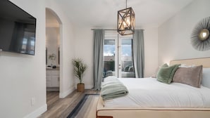 Ein Schlafzimmer mit Bett, einem an der Wand befestigten Fernseher, einem Blumentopf und Blick durch eine Glastür.