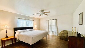 Un dormitorio con cama, dos lámparas de noche, ventilador de techo, ventana con cortinas, piso de madera y cómoda decorada.