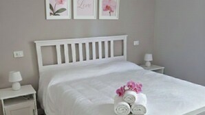 Ein ordentlich bezogenes Bett mit weißer Bettwäsche und rosa Blumen, zwei Nachttische mit Lampen sowie drei gerahmte Bilder an der Wand.