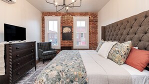 Un dormitorio con pared de ladrillo, una cama grande con muchas almohadas, un televisor sobre una cómoda y una silla.