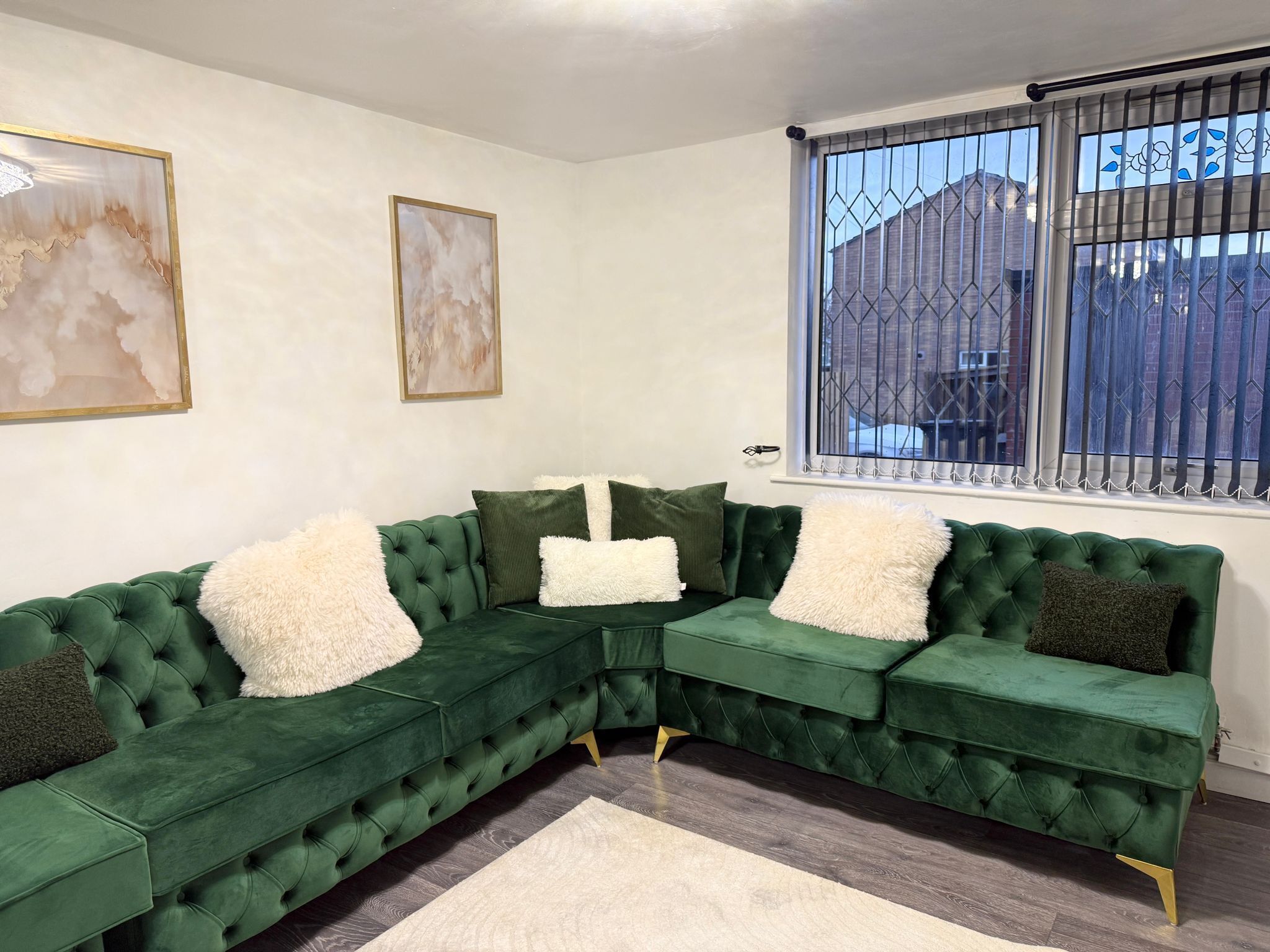 Un sofá seccional verde con cojines blancos y verde oscuro, una alfombra blanca y dos cuadros abstractos en la pared.