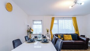 Un comedor con una mesa blanca dispuesta para cuatro personas, un sofá de cuero marrón con cojines, y un reloj amarillo en la pared.