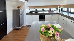 Een moderne keuken met een eethoek, met een centraal kookeiland met bloemenarrangement, een inbouwoven en een groot raam dat natuurlijk licht binnenlaat.