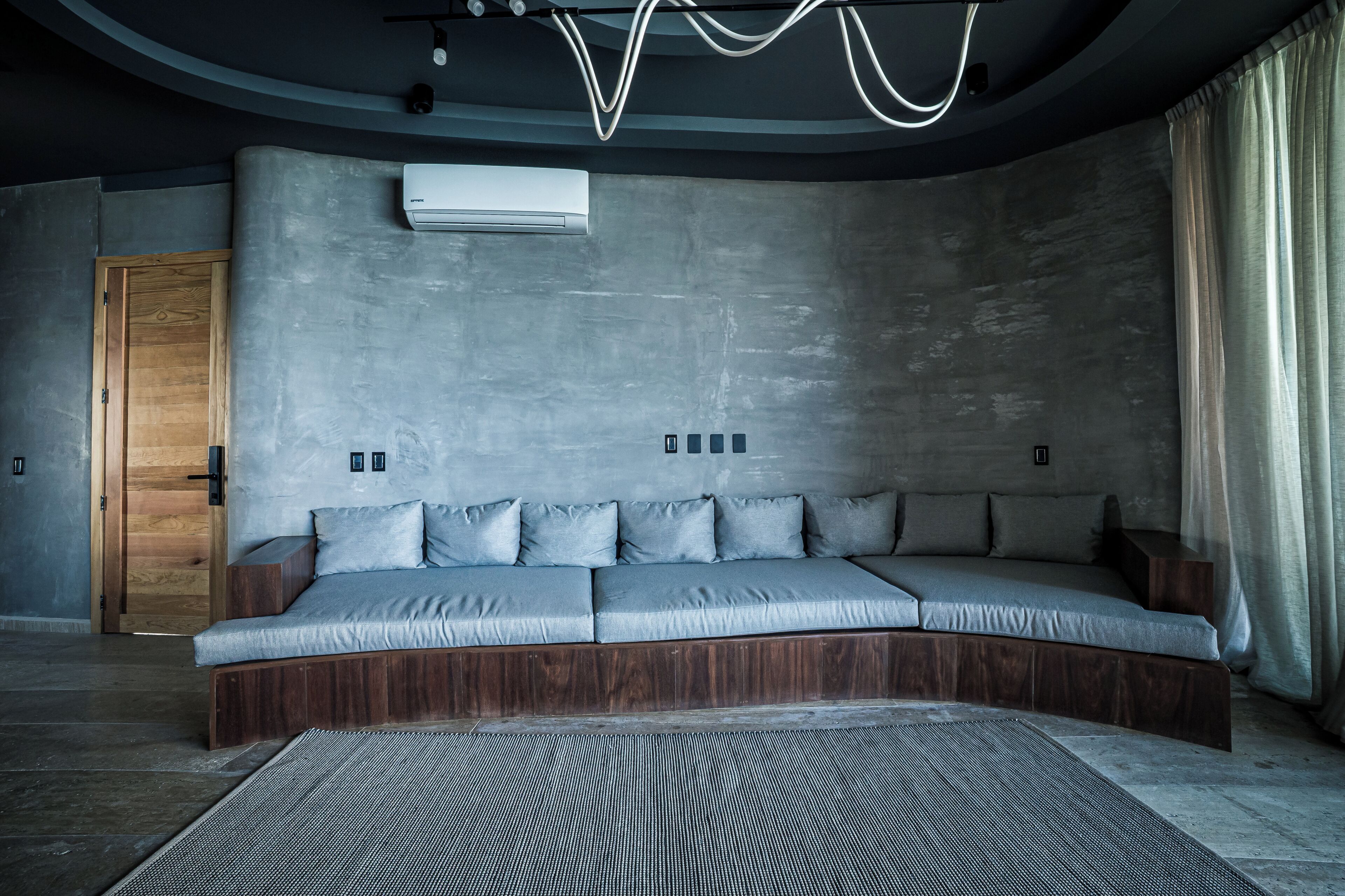 Un canapé-lit moderne au design incurvé, doté d’une base en bois et de coussins gris, dans une pièce avec un mur en béton, une porte en bois et une grande fenêtre agrémentée de rideaux.