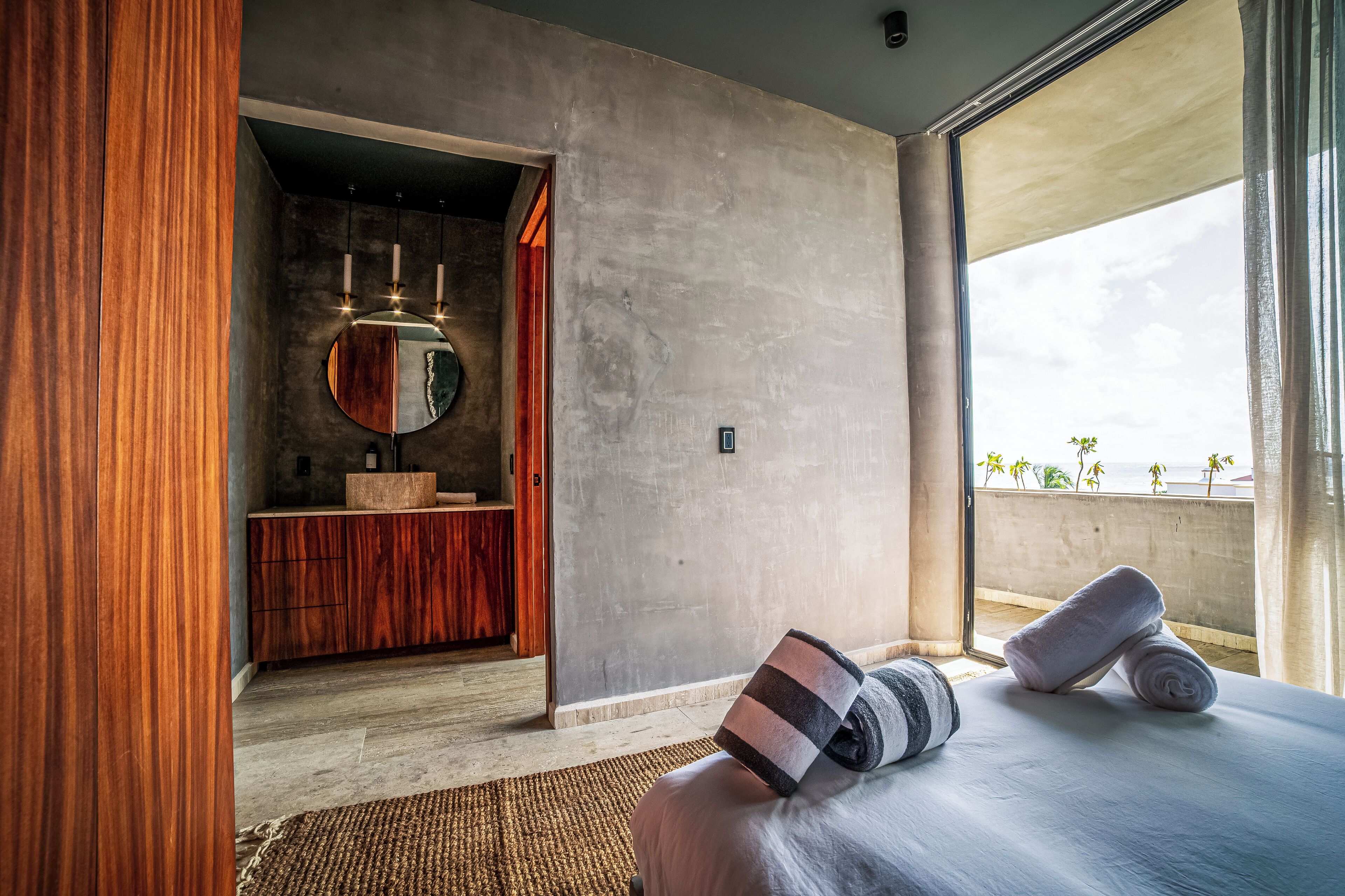 A modern bedroom with a large bed, a wooden vanity, a round mirror, and a view of the outdoors.