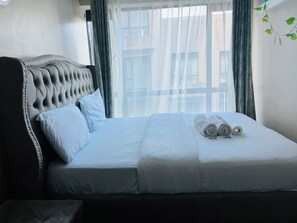A neatly made bed with white linens and rolled towels, a tufted headboard, and sheer curtains allowing natural light. - Clean Modern 1BR near TRM Mall off Thika Road
Perfect for long and Short stays. (Nairobi)