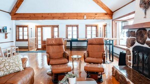 A spacious living room with high ceilings, wooden beams, a fireplace, and a view of the kitchen through large windows. - Le Repere du Lac - Lake View, Access, beach & Spa (Venise-en-Québec)