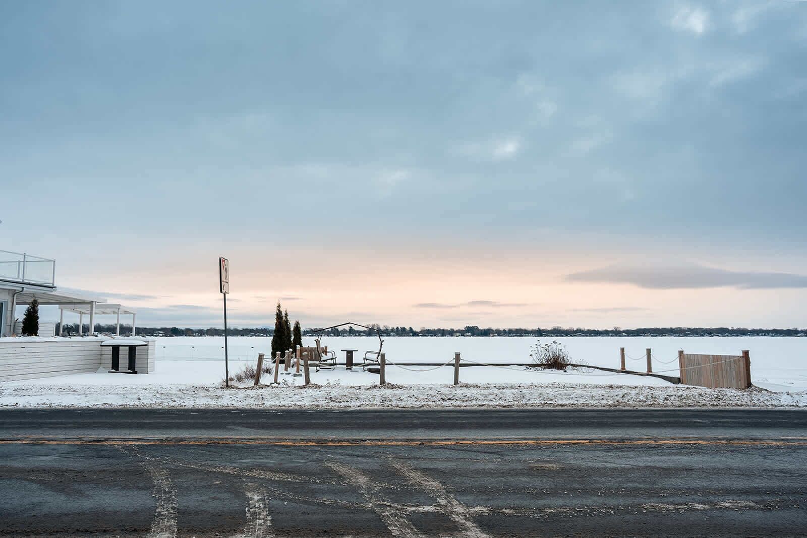 A snowy landscape with a building on the left, a wooden fence, and a body of water in the background.
