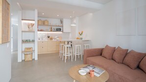 A modern living room with a brown sofa, a round coffee table, and a kitchen area with white bar stools and a white countertop. - With patio, in Funchal city centre - AL'gria I (Funchal)