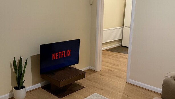 A living room with a flat-screen TV displaying 'NETFLIX', a potted plant, and two beige sofas. - 3-Bed Home, Free Parking, Smart TV,Family & Business Ready (Slough)