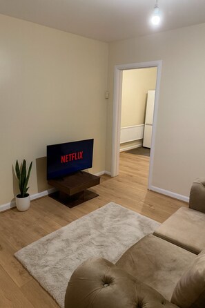 Un salon avec un téléviseur à écran plat affichant le logo « NETFLIX », une plante en pot et deux canapés de couleur beige.