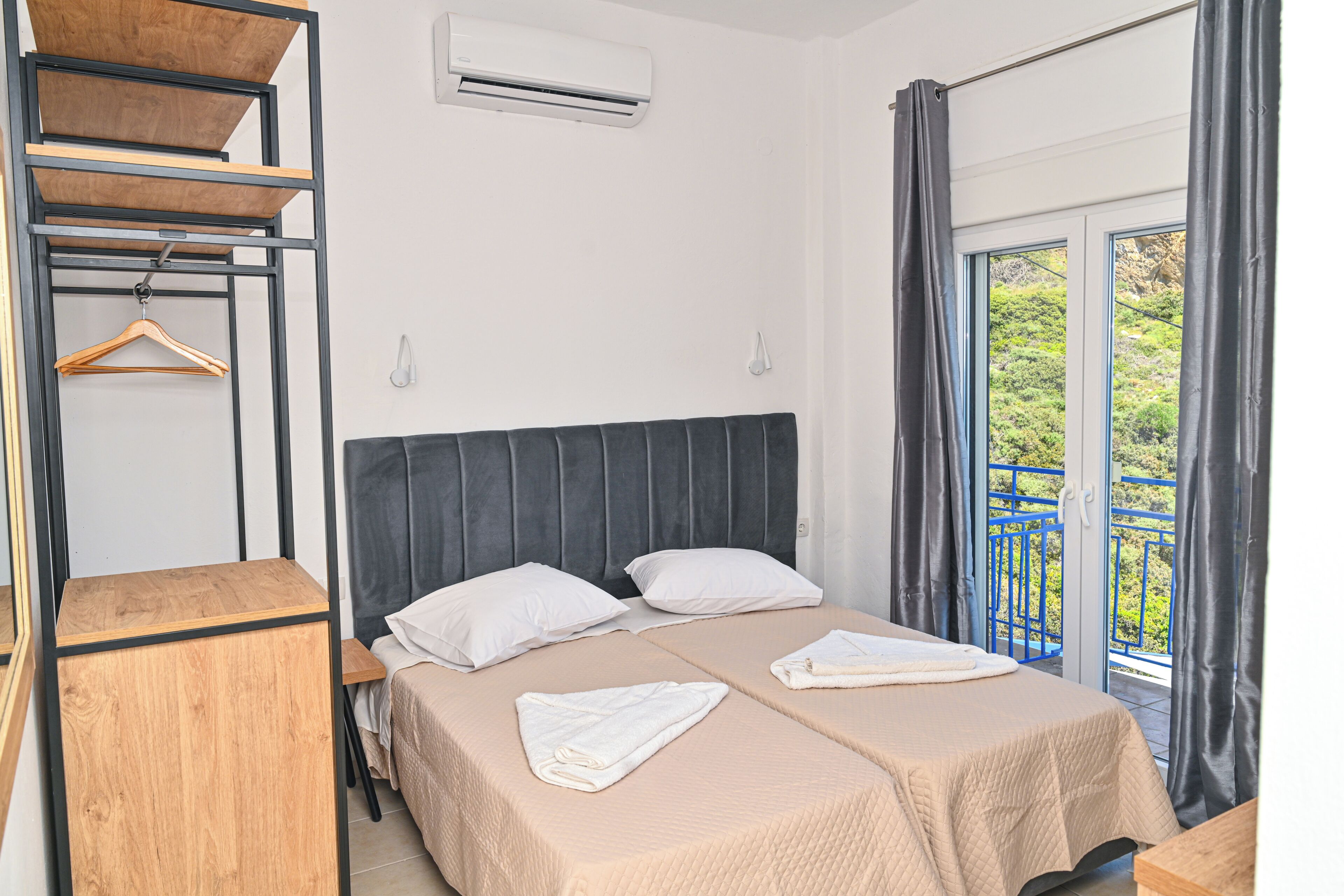 A double bed with a headboard, two white pillows, and a beige bedspread. There is a wooden side table, a hanging wooden hanger, and a window with curtains.
