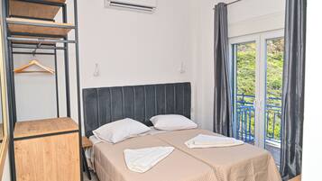 Una cama doble con cabecera, dos almohadas blancas y una colcha color beige. Hay una mesita de noche de madera, un perchero de madera colgante y una ventana con cortinas.