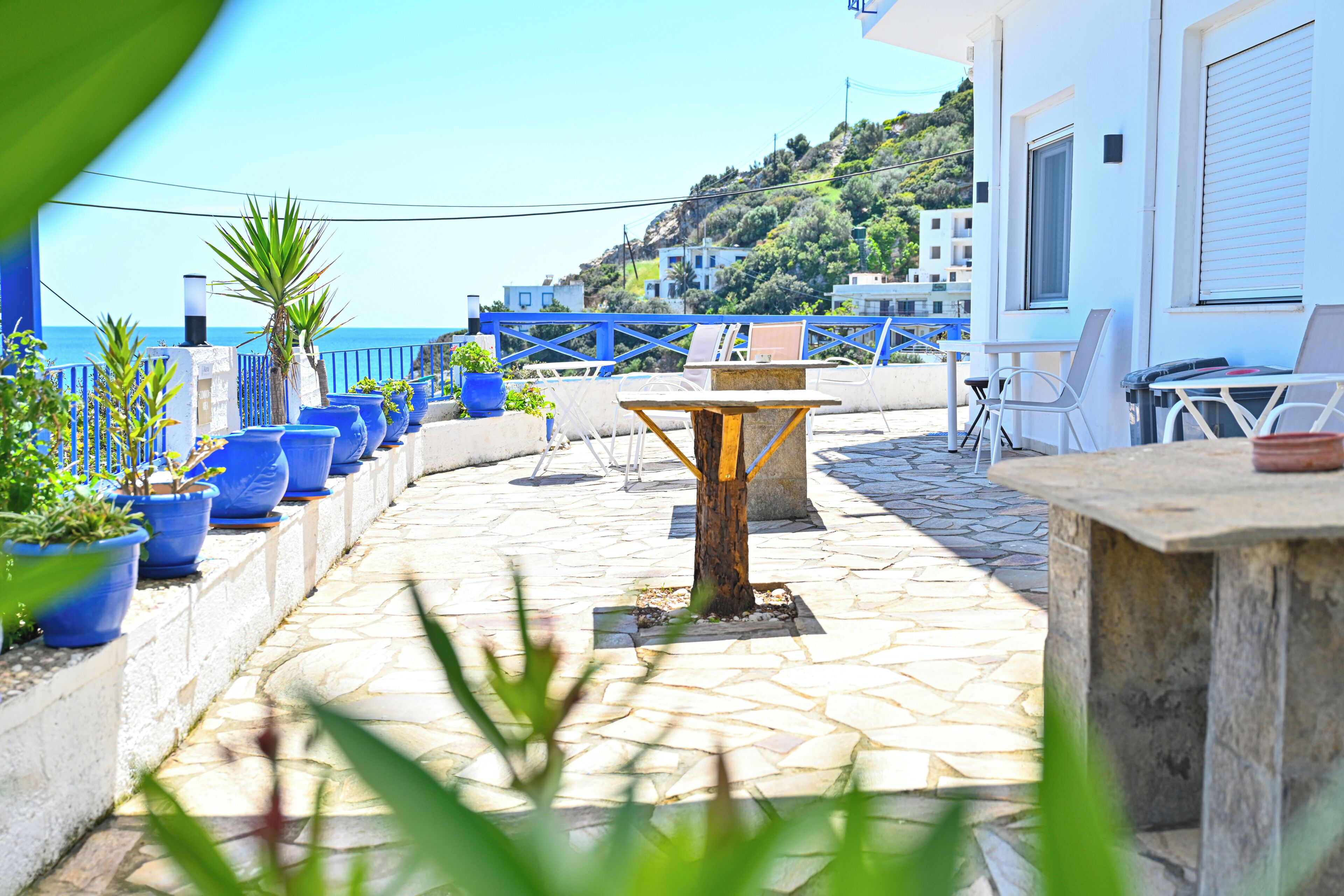 A terrace with potted plants, a table, and chairs, overlooking a coastal area with buildings and hills.