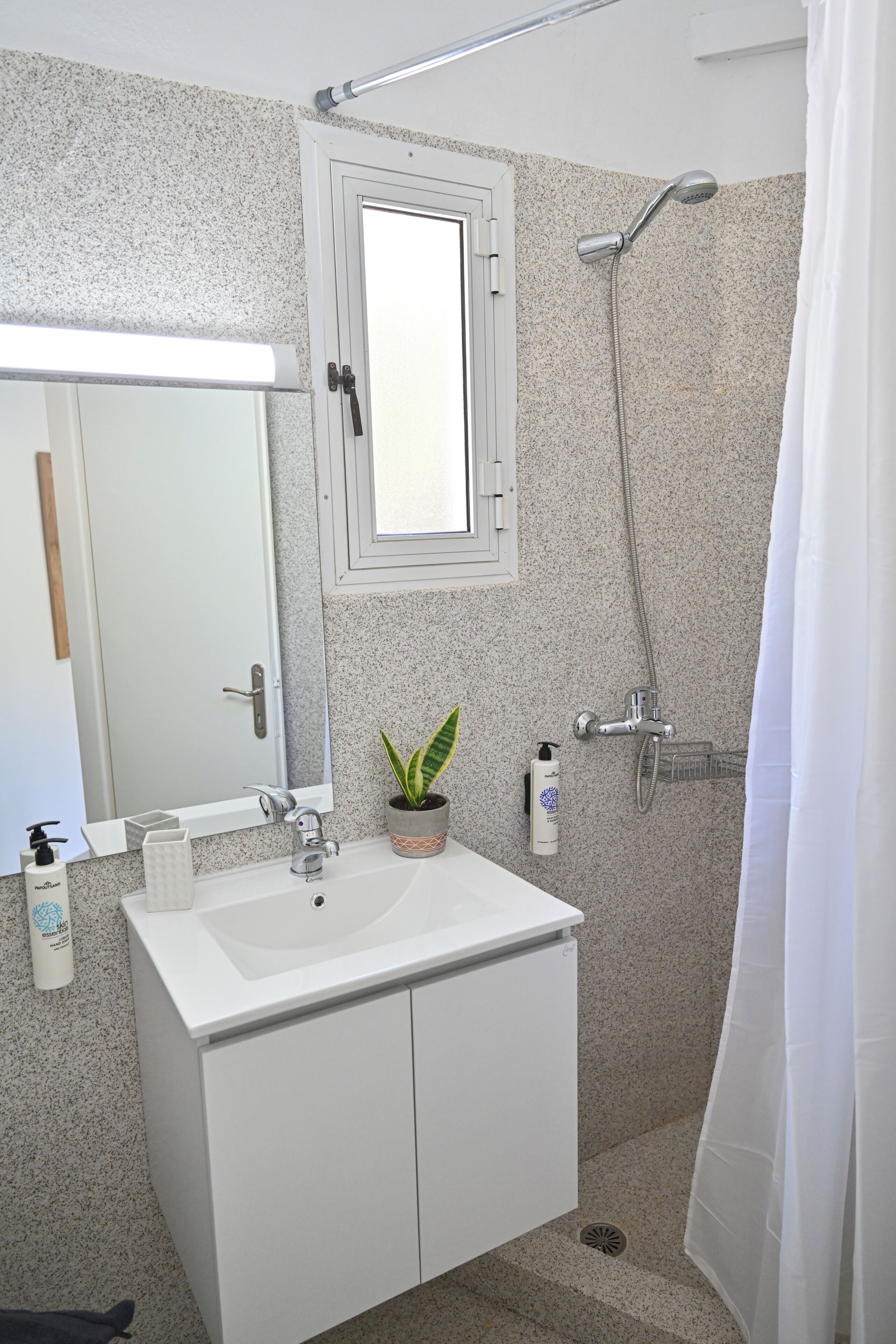 Un baño con un lavamanos blanco, un espejo, una ducha y una planta en maceta.