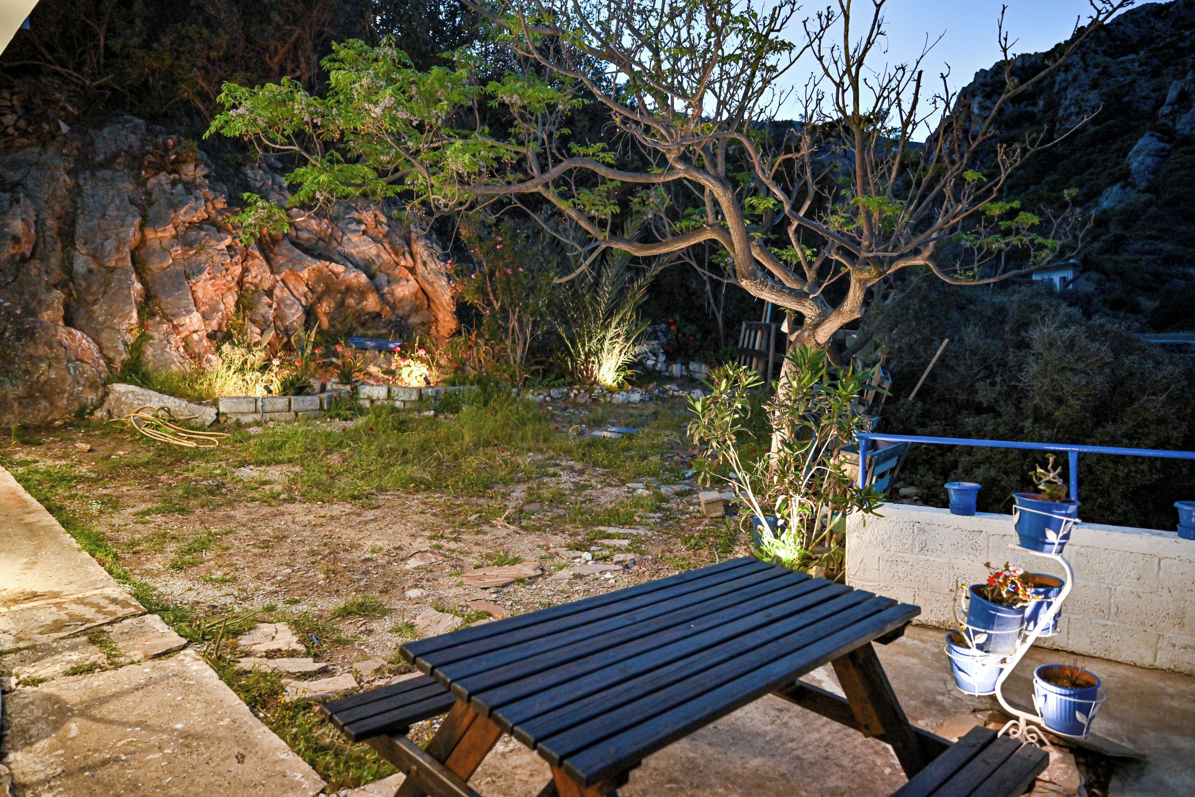 A patio area with a wooden table and benches, surrounded by potted plants and a rocky landscape.