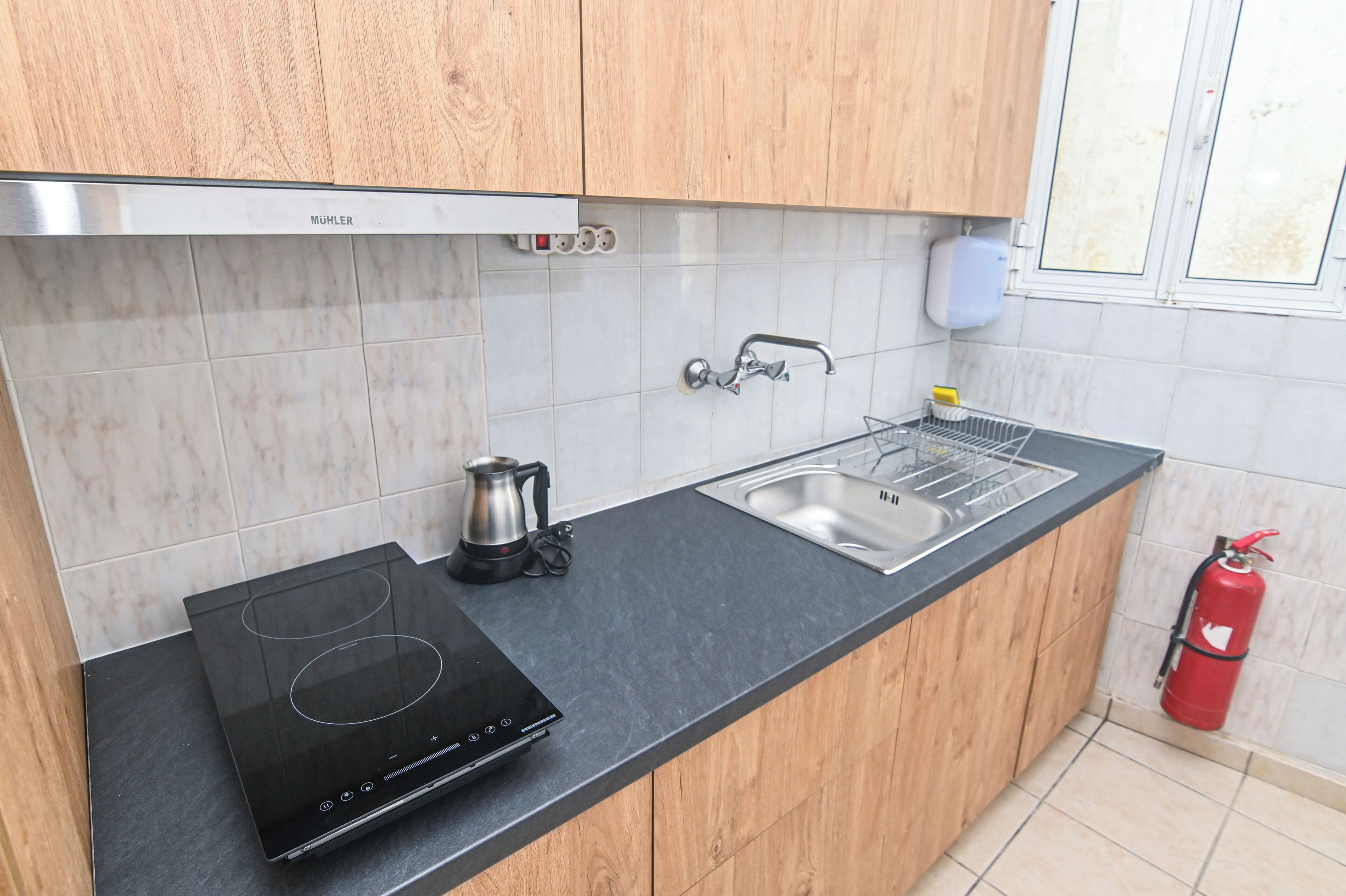 A kitchen with a black induction cooktop, a stainless steel sink, wooden cabinets, and a red fire extinguisher.