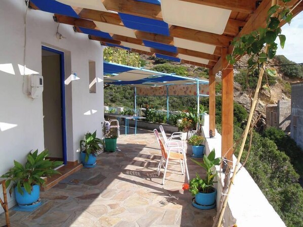 A covered patio area with stone flooring, outdoor furniture, and potted plants. - IKARIA THERMA BLUE (Ikaria)