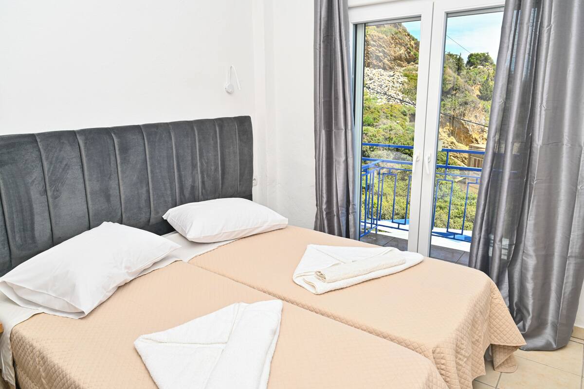 Ein Hotelzimmer mit Bett, Kissen, einem Fenster mit Vorhängen und Blick auf Grünfläche.