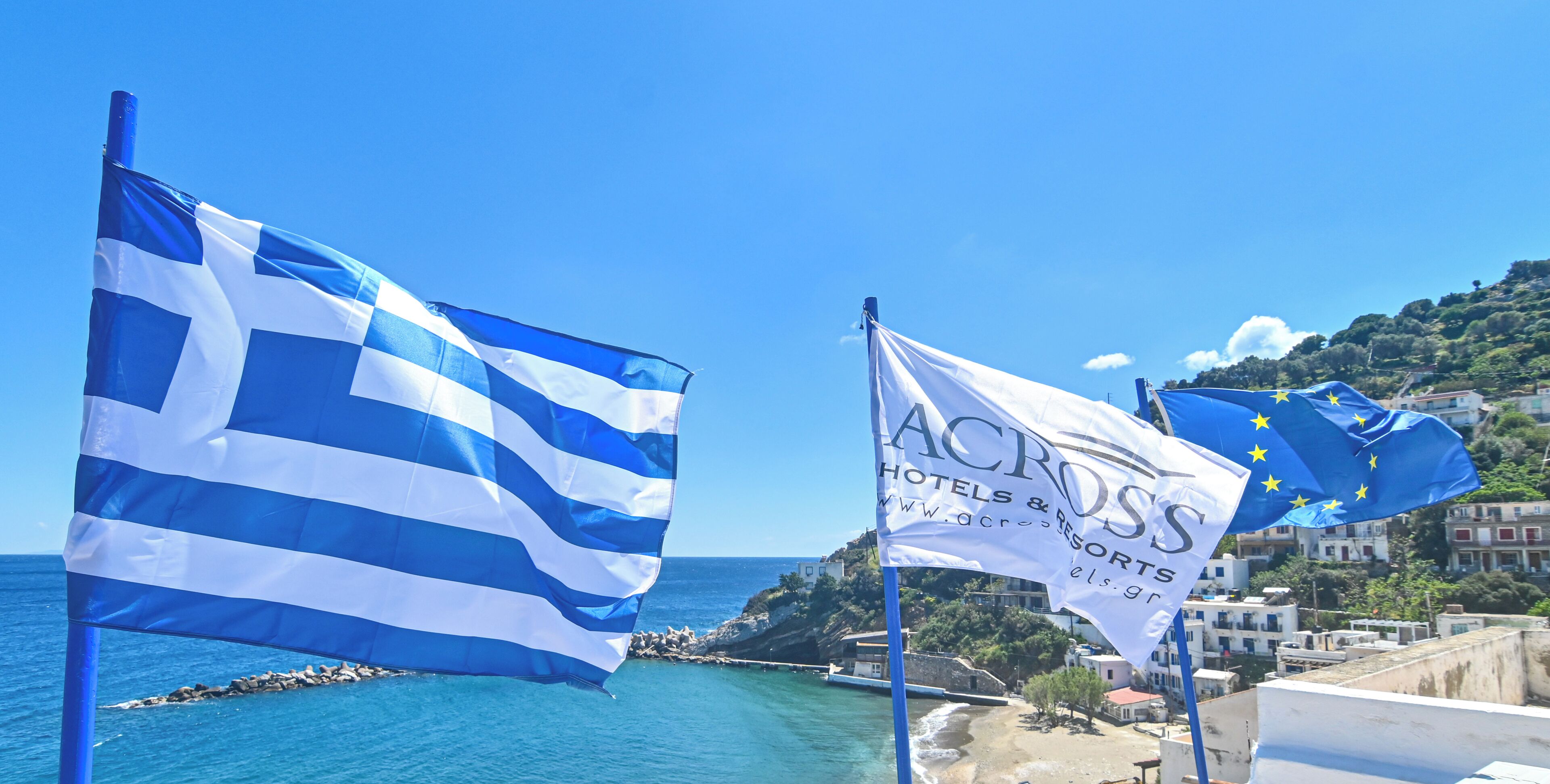 Dos banderas, una griega y otra de la Unión Europea, con un pueblo costero al fondo.