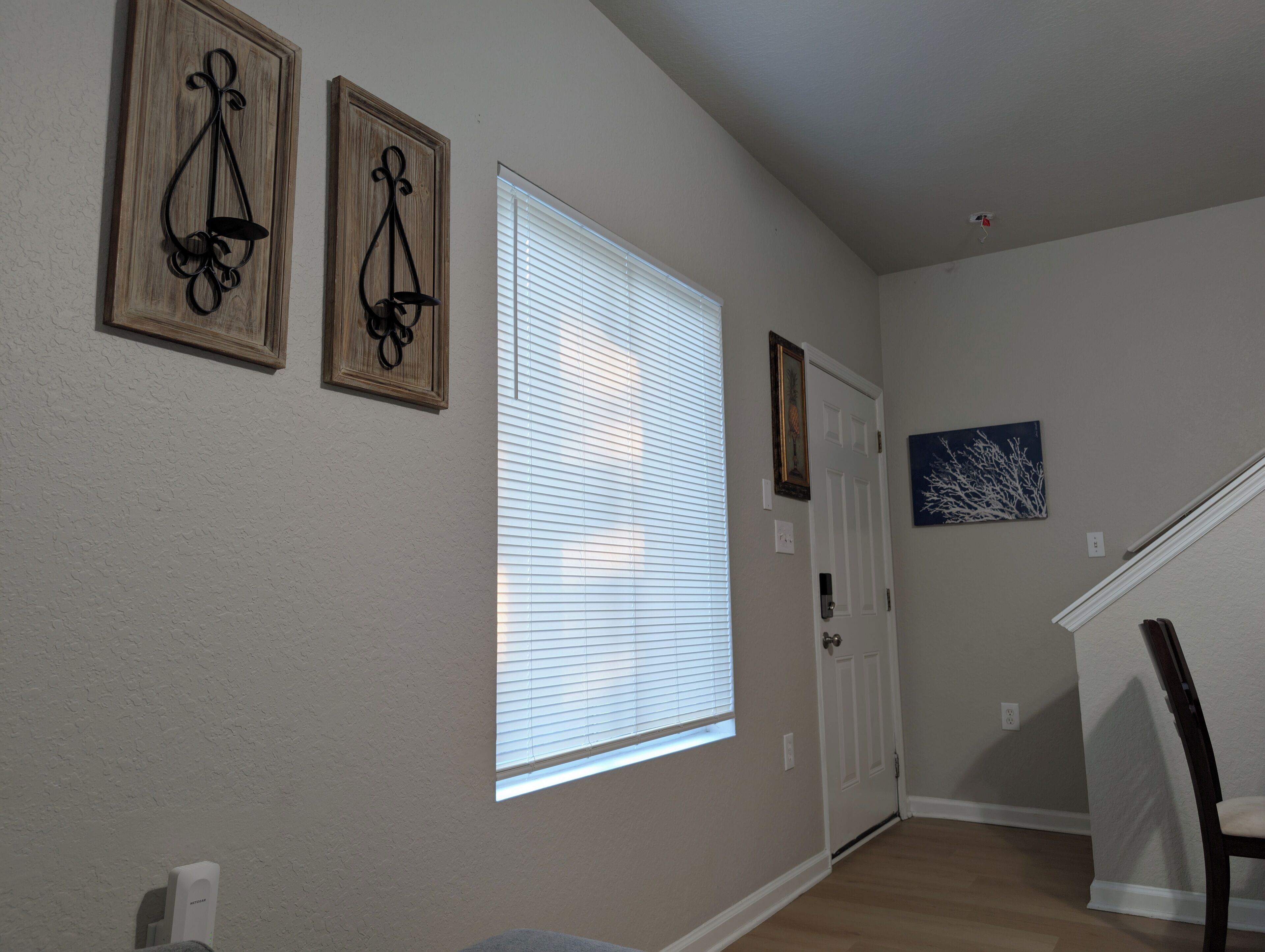 A hallway with a wooden floor, a white door, and a window with blinds. There are two wall-mounted decorations and a painting on the wall.