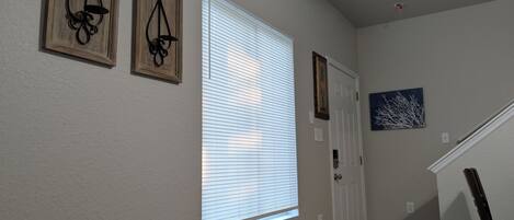 A hallway with a wooden floor, a white door, and a window with blinds. There are two wall-mounted decorations and a painting on the wall.