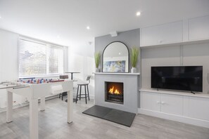 A modern living room with a foosball table, a fireplace, a television, and a large window with blinds. - Fantastic 3 Bedroom Home in Gloucester City (Gloucetser)
