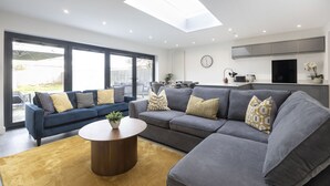 Ein modernes Wohnzimmer mit einem grauen Ecksofa, einem runden Couchtisch und einem großen Fenster mit Blick ins Freie.