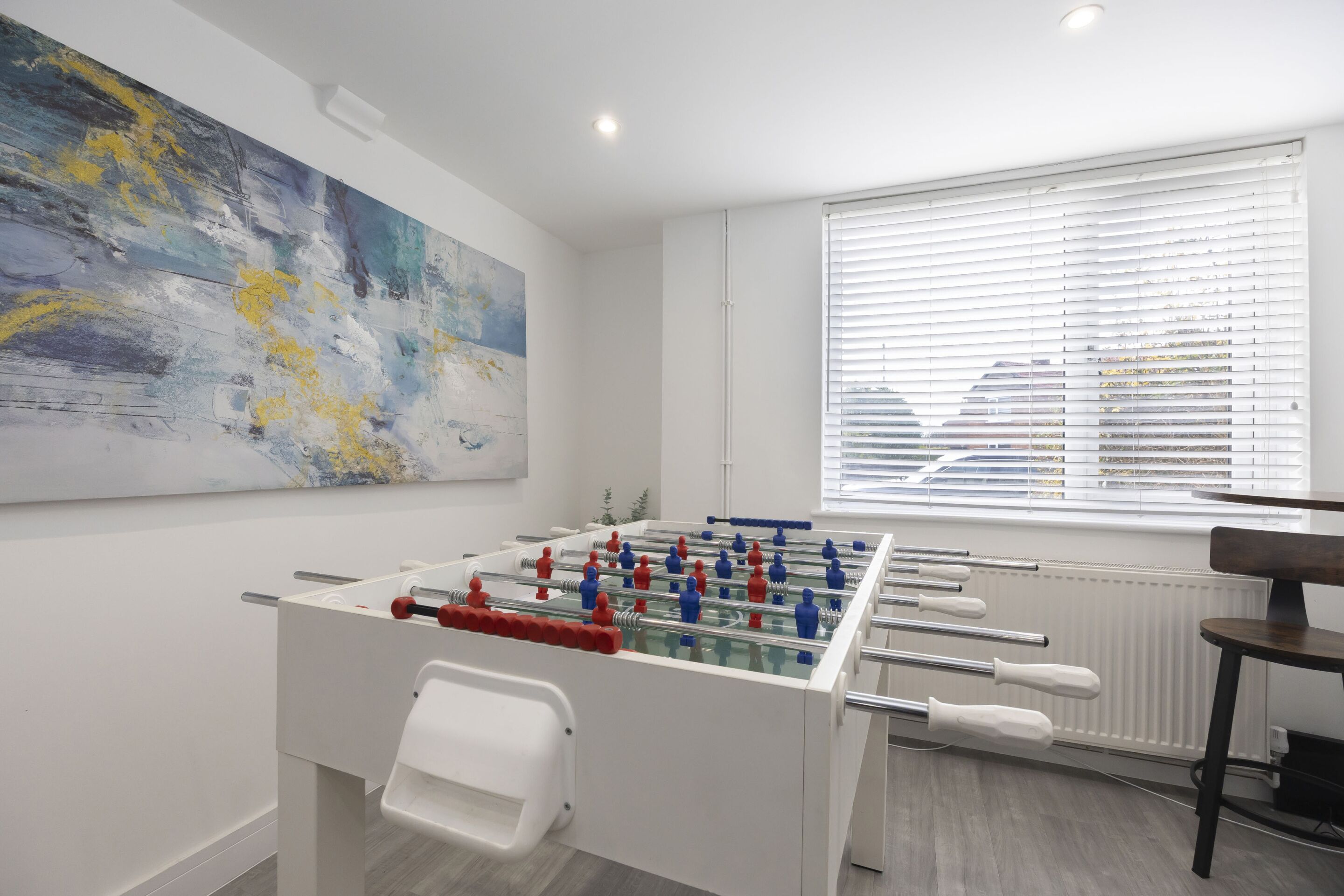 Een moderne kamer met een tafelvoetbaltafel, een groot abstract schilderij en een raam met luxaflex.