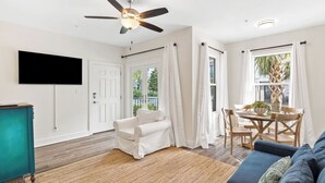 A living room with a TV, ceiling fan, white sofa, dining table, and a view of greenery outside. - Chic condo@Hidden Lake~Santa Rosa Beach ~Sleeps 6! (Santa Rosa Beach)