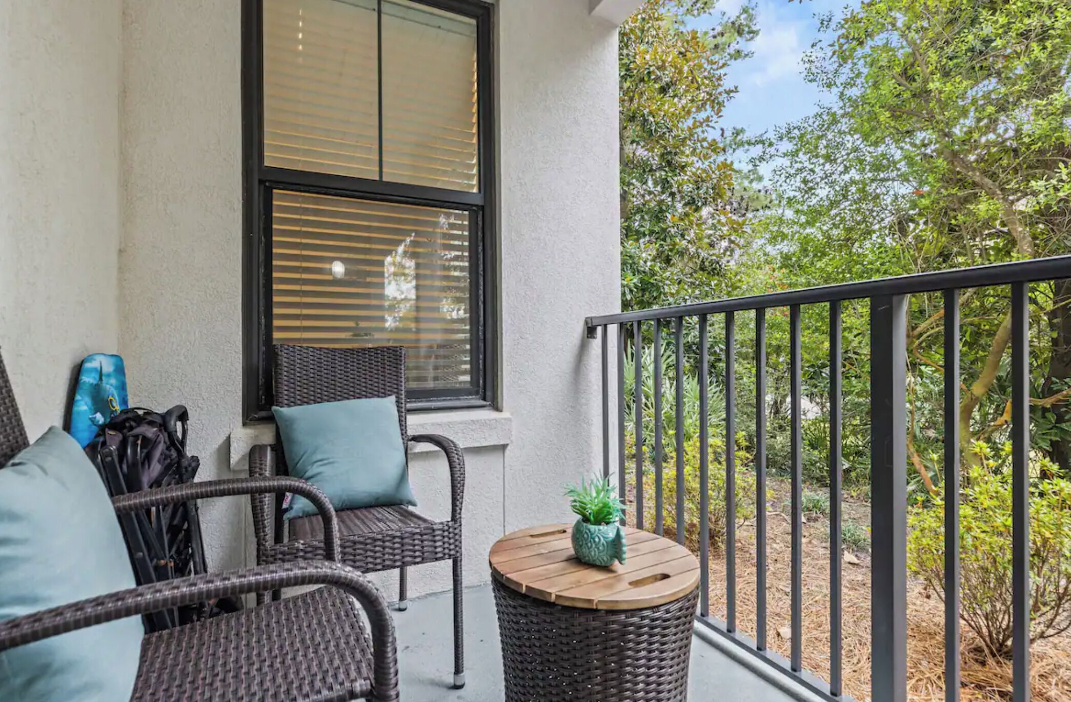 A balcony with wicker furniture, a small round table with a plant, and a view of trees and shrubs.