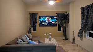 Ein Wohnzimmer mit einer Couch, einem an der Wand montierten Fernseher, einem Deckenventilator und Vorhängen.
