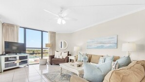 A spacious living room with a large flat-screen TV, a ceiling fan, and a view of the outdoors through large windows. - 3 Bd/3Ba GULF views! Comp Beach chair set-up! (Miramar Beach)