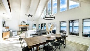 Amplia sala de estar con chimenea, mesa de comedor y grandes ventanales con vista al agua.