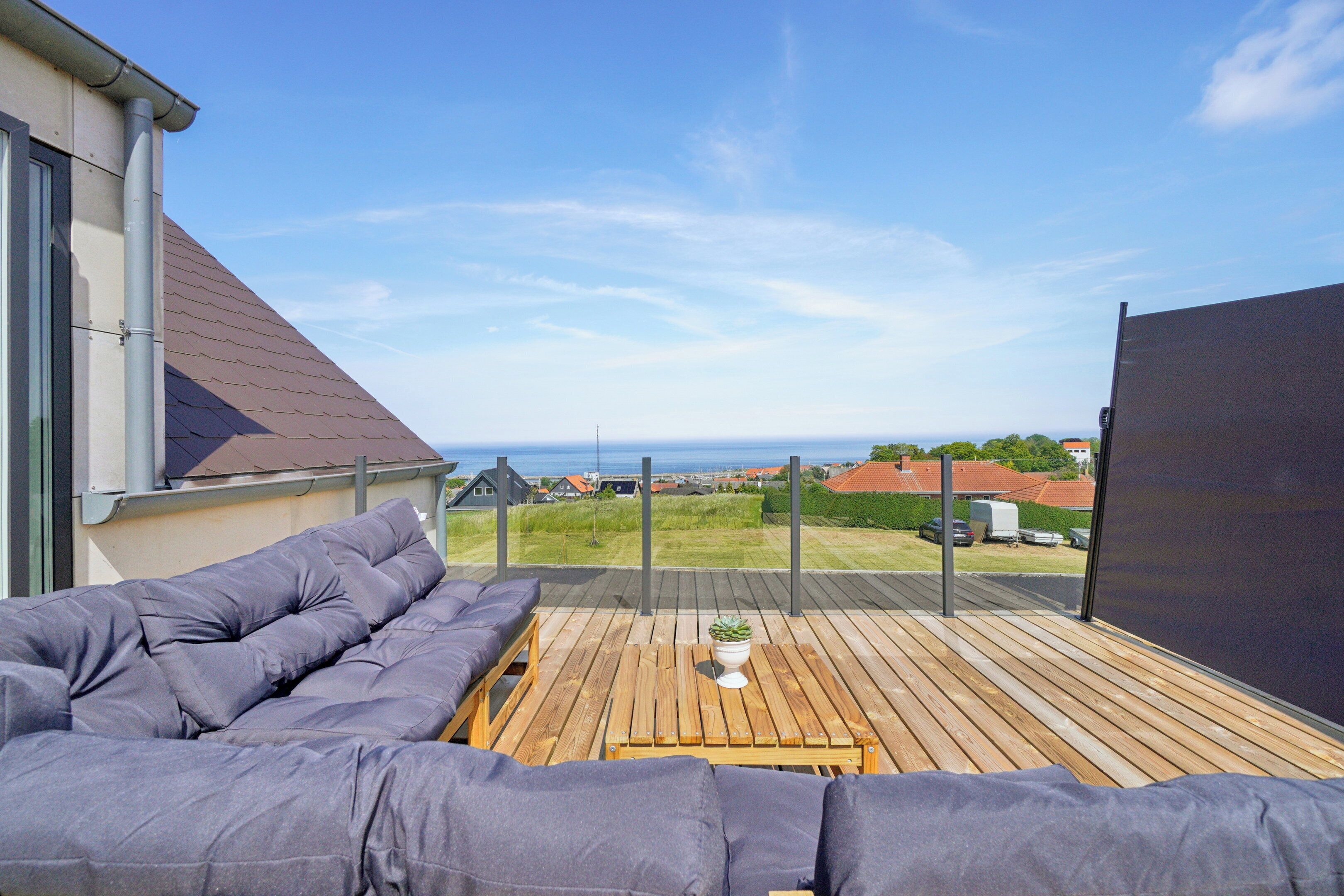 Een houten terras met een bank en een tafel, met uitzicht op een woonwijk en de zee.